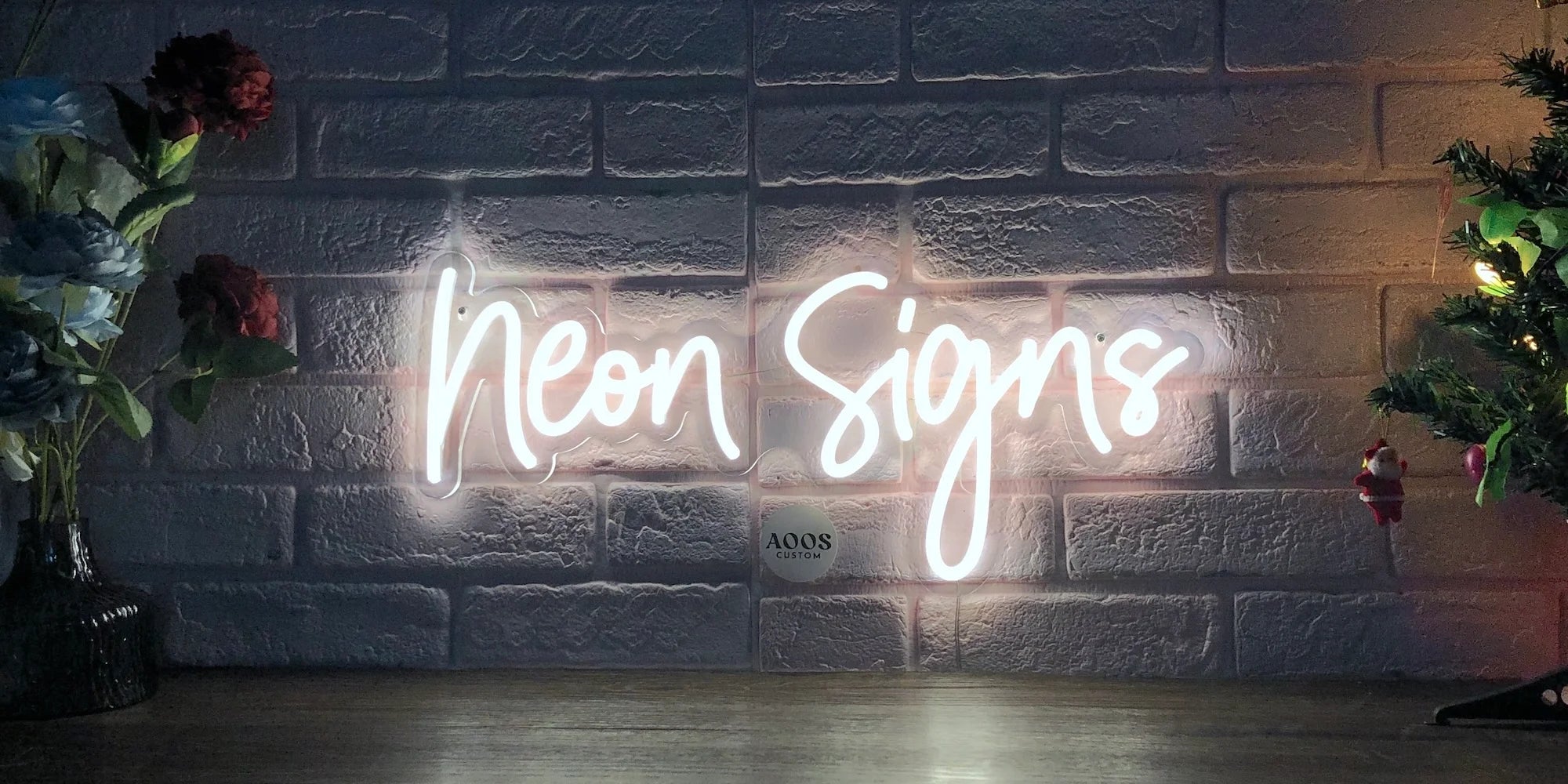 A white LED neon sign spelling "Neon Signs" mounted on a transparent acrylic board against a textured brick wall, with decorative flowers and a Christmas tree nearby.
