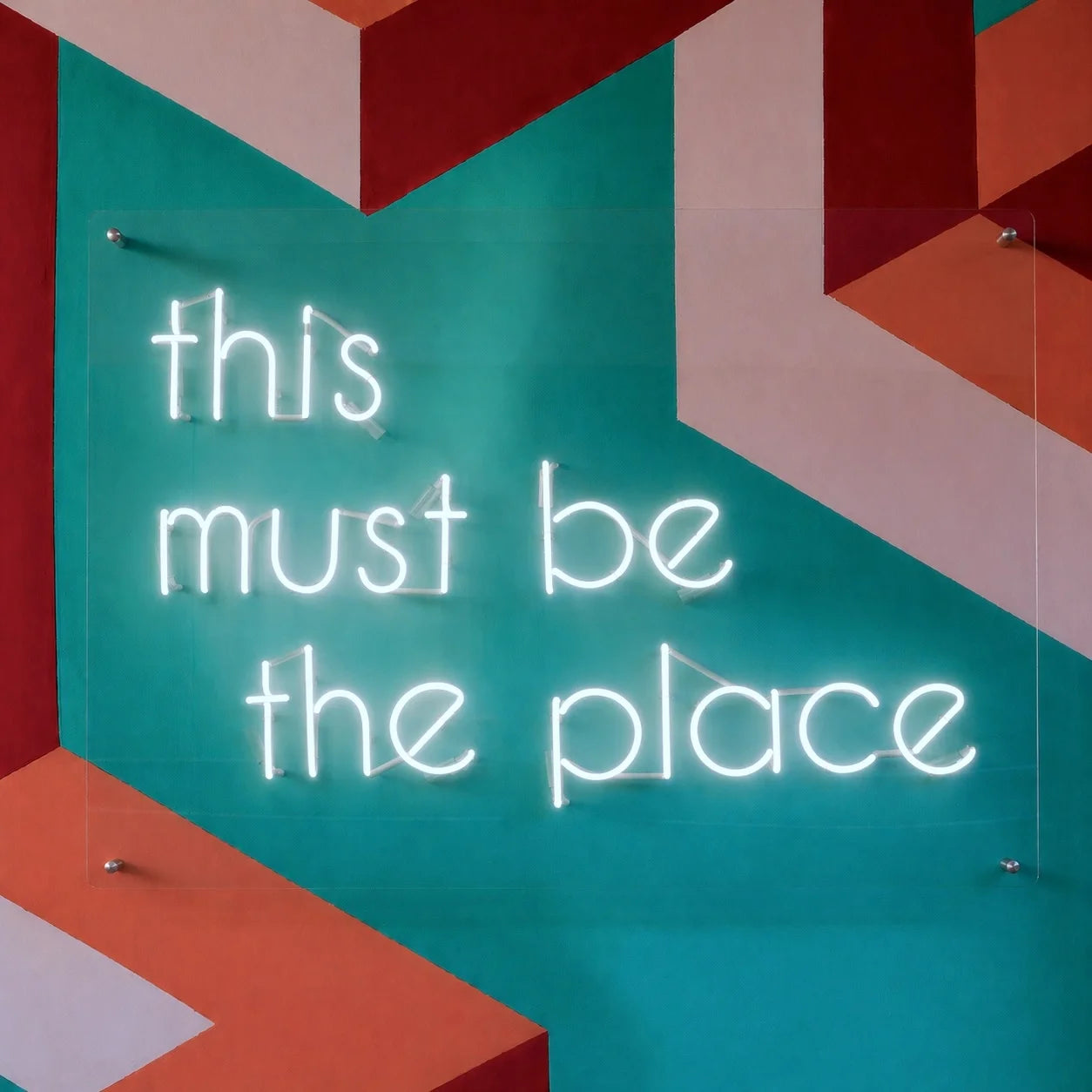 Custom real glass neon sign with personalized text or logo formed from hand bent glass tubing. The sign uses traditional neon illumination with colored glass tubes mounted on a clear acrylic backing panel or metal support structure for wall installation. The design can include script lettering, block text, or logo outlines with a continuous illuminated tube line. The sign is suitable for storefront branding, bars, salons, restaurants, studios, retail displays, and interior business walls.