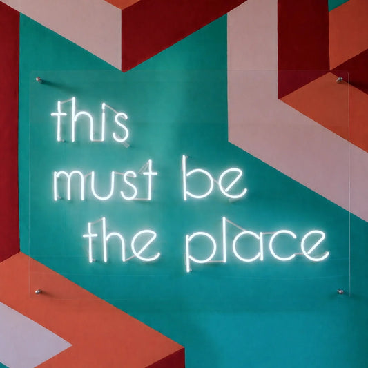 Custom real glass neon sign with white phrase lettering on clear acrylic backing mounted on a colorful geometric wall