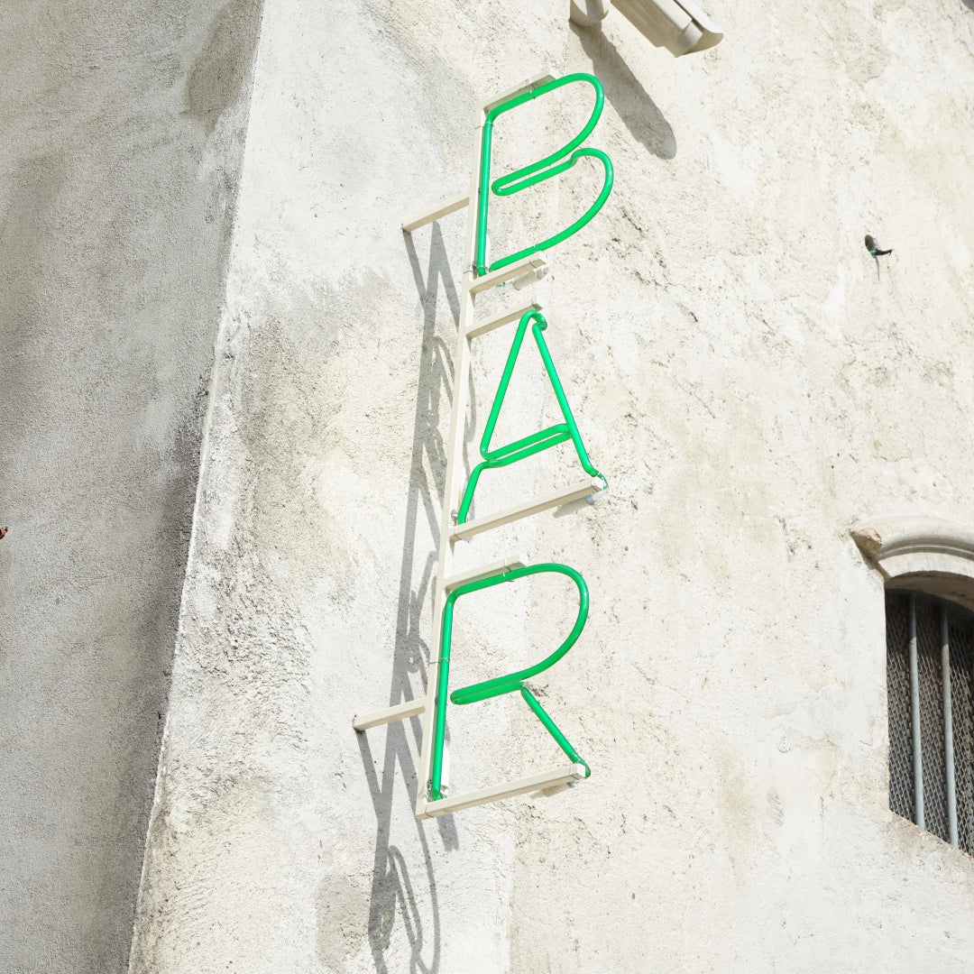 Custom real glass neon art sign with green BAR lettering in a vertical blade sign format mounted on an exterior stucco wall