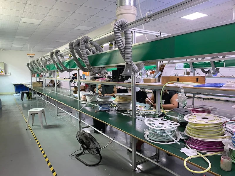Large workshop with an assembly line for neon signs, featuring workers, green workstations, exhaust ducts, and reels of neon tubing.